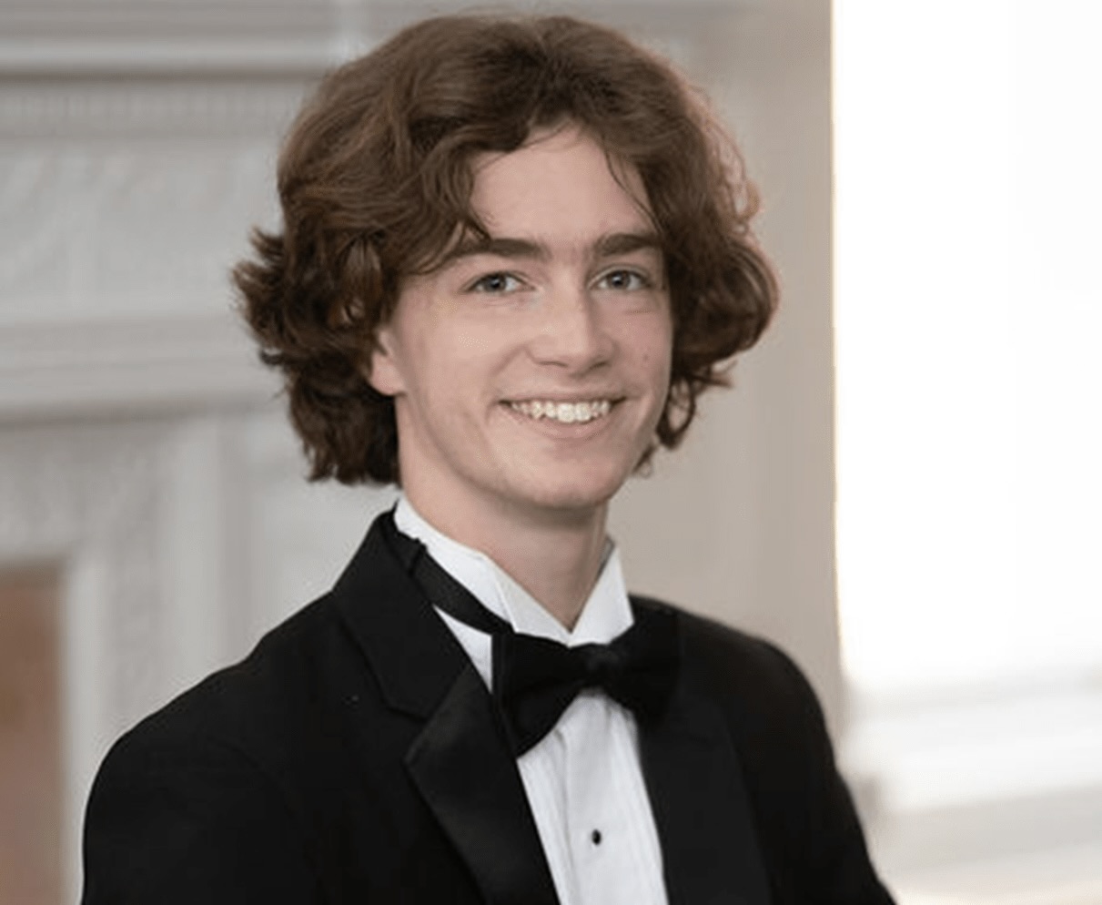 A young man sits smiling by a piano